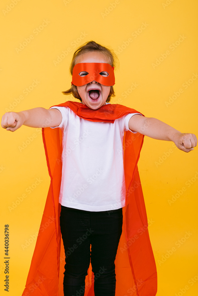 Little blonde girl dressed as a superheroine superhero with cape and red mask, screaming with open arms in flying position, on yellow background