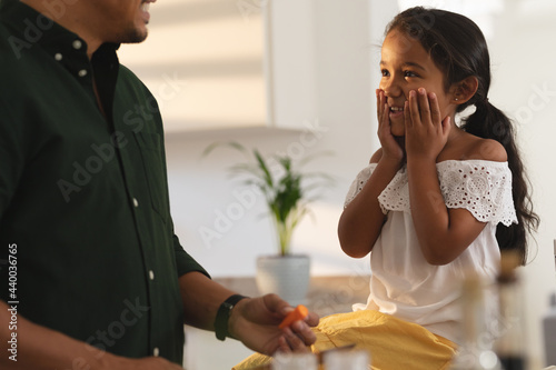 Happy hispanic daughter sitting on counter making face to father preparing vegetables in kitchen