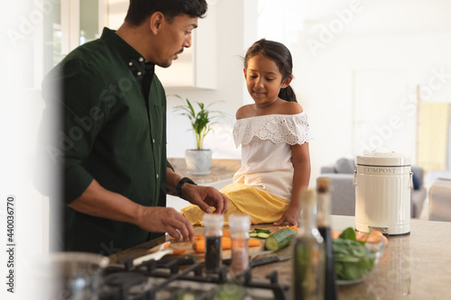 Happy hispanic daughter and father preparing vegetables in kitchen, daughter sitting on counter photo