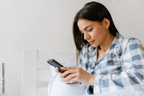 Young hispanic woman using cellphone while lying in bed at home