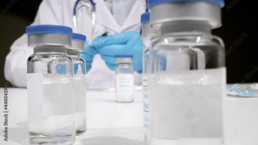 Sliding Shot Approaching a Covid 19 Vaccine Vial as Medical Professional In Background Prepares to Administer It Sliding Shot Approaching a Covid 19 Vaccine Vial as Medical Professional In Background Prepares to Administer It