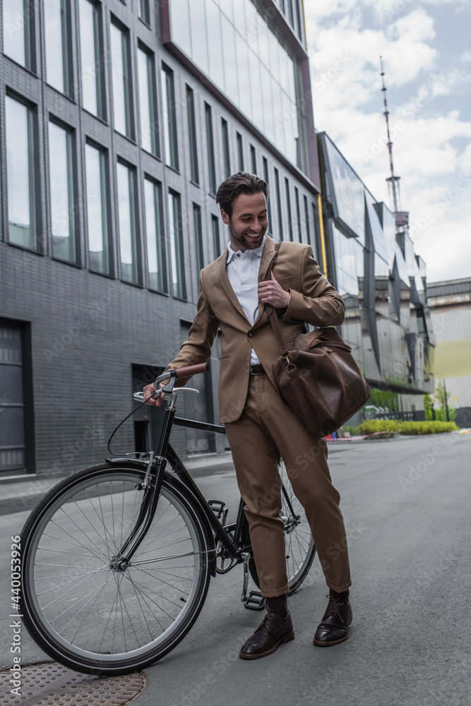 Obraz premium full length of happy businessman in suit holding leather bag and smiling near bicycle outside.