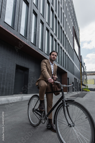 full length of young businessman in suit with leather bag riding bicycle outside.