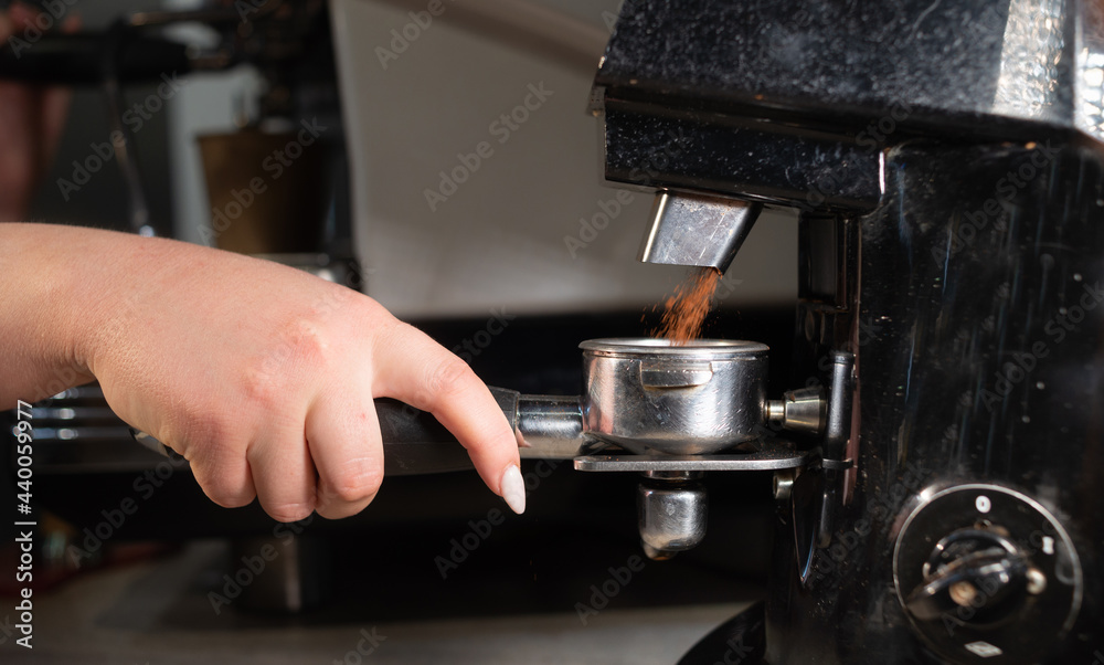Barista grinds coffee in a professional coffee grinder holding a chrome portafilter in his hand. Preparing grain for making espresso in a cafe. Close-up