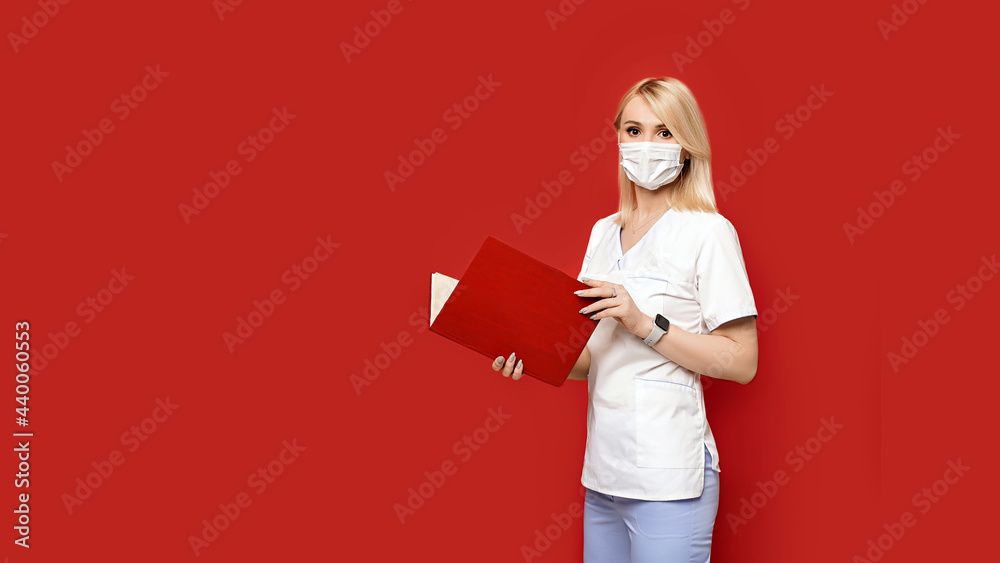 Confident female doctor wearing protective face mask standing on red background with notebook or medical book in her hands. Young woman intern in medical clothes posing.