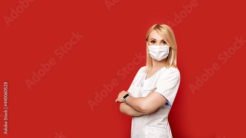 Obraz na płótnie Confident female doctor wearing protective face mask standing on red background arms crossed. Young woman intern in medical clothes posing. w salonie