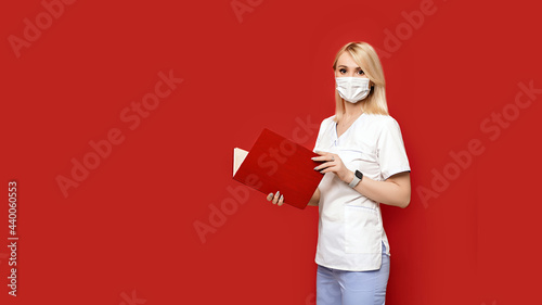 Obraz na płótnie Confident female doctor wearing protective face mask standing on red background with notebook or medical book in her hands. Young woman intern in medical clothes posing. w salonie
