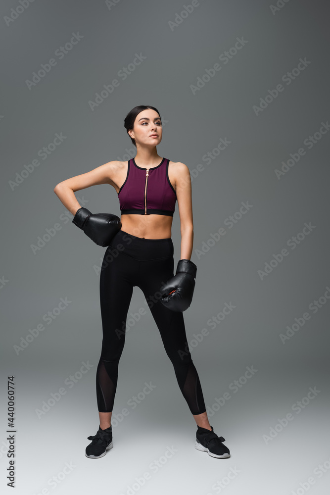 confident sportswoman in boxing gloves standing with hand on hip on grey background.