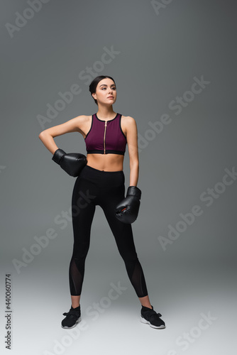 confident sportswoman in boxing gloves standing with hand on hip on grey background.