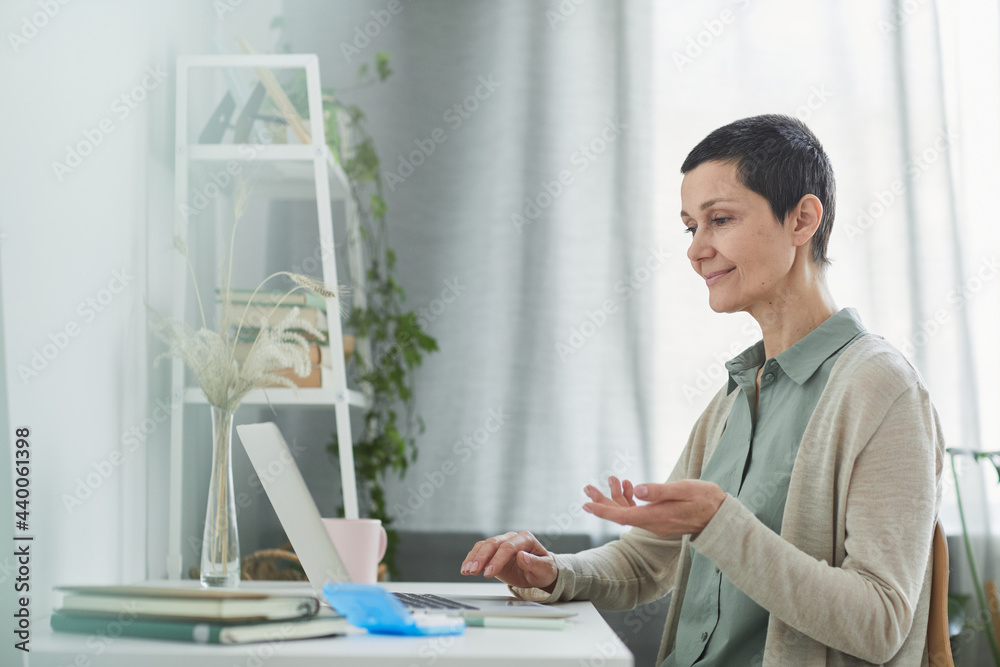 Side view at smiling mature woman speaking by video call and using laptop in cozy home setting, copy space Side view at smiling mature woman speaking by video call and using laptop in cozy home setting, copy space