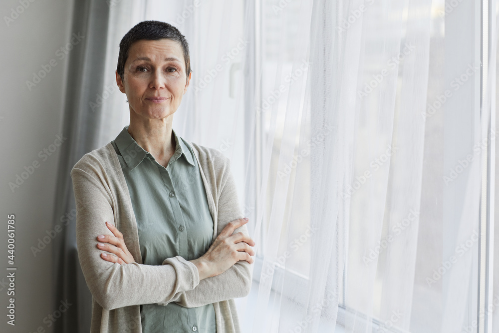 Waist up portrait of mature woman looking at camera while standing by window with arms crossed, copy space Waist up portrait of mature woman looking at camera while standing by window with arms crossed, copy space
