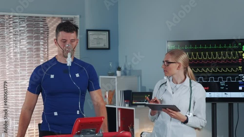 Athlete in oxygen mask doing stress test while cardiologist with tablet monitoring his EKG on display in modern scientific sports laboratory