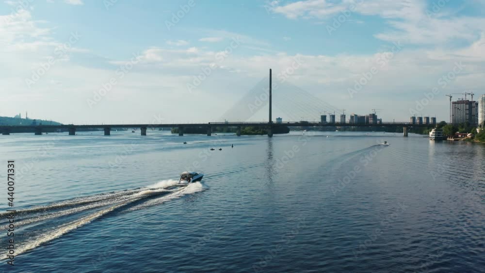 Aerial view of motorboat sailing on wide river against background of large bridge and cityscape. Drone camera flying over water behind high-speed powerboat making waves at daylight Aerial view of motorboat sailing on wide river against background of large bridge and cityscape. Drone camera flying over water behind high-speed powerboat making waves at daylight