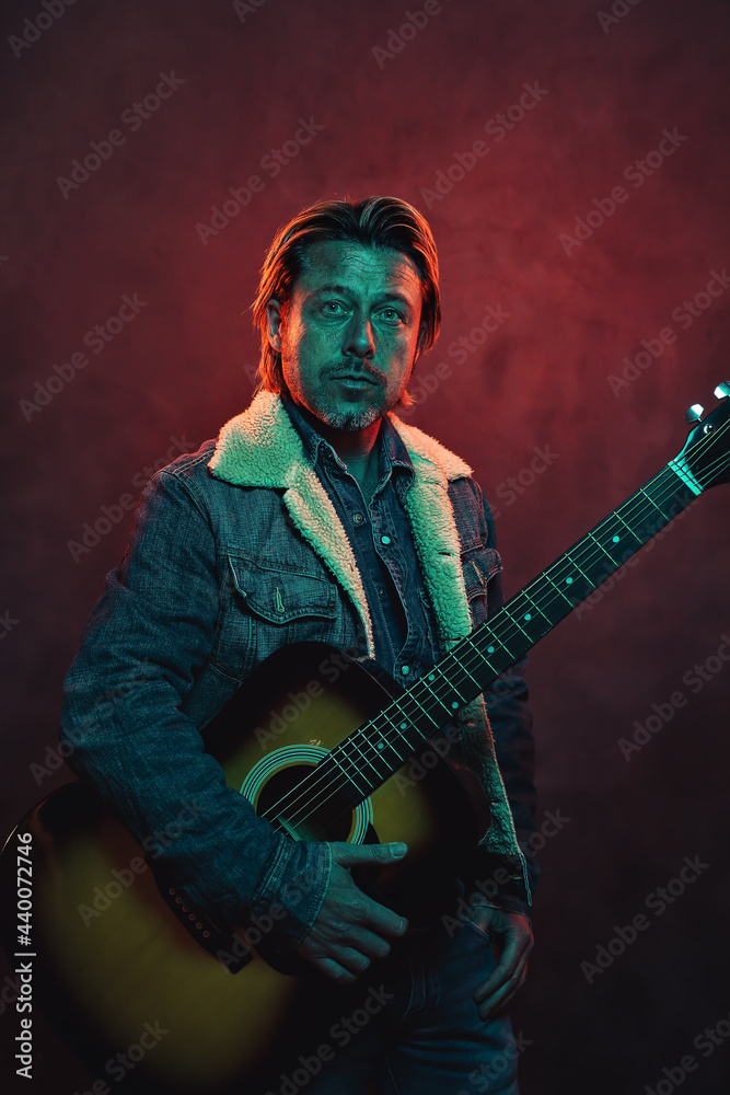 Man with acoustic western guitar in jeans jacket in red and blue colored light.