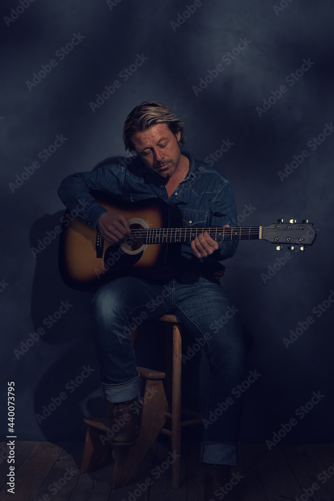 Obraz premium Man in denim shirt and jeans with acoustic western guitar on a wooden stool in dappled sunlight in front of a grey wall.