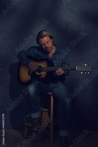 Man in denim shirt and jeans with acoustic western guitar on a wooden stool in dappled sunlight in front of a grey wall.