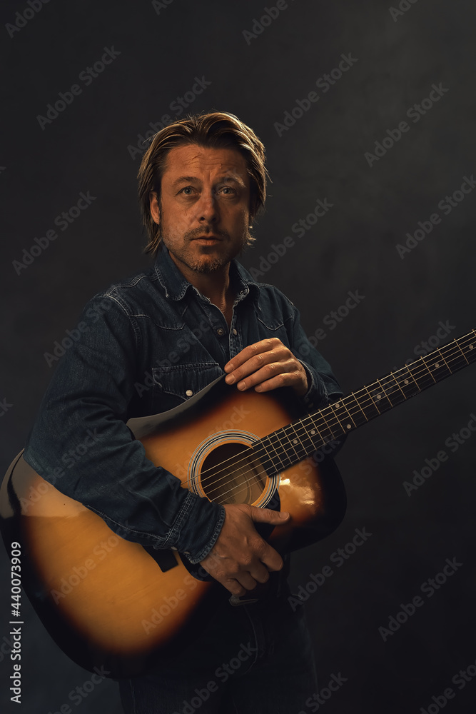 Man in denim shirt and jeans with acoustic western guitar in front of a grey wall.