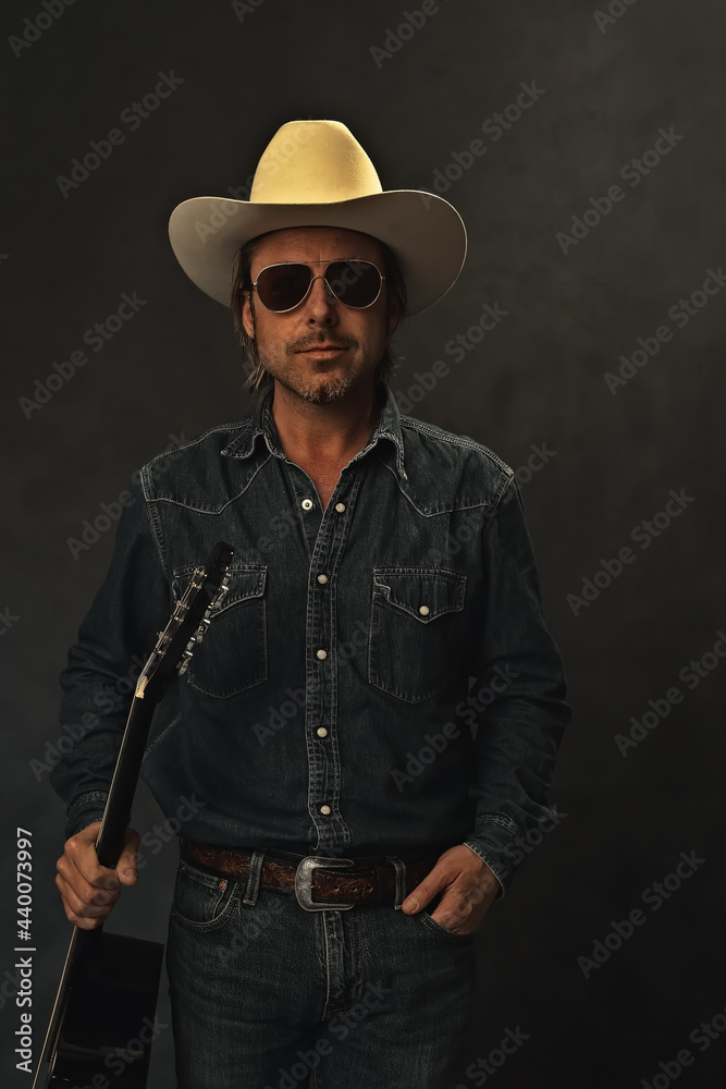 Man in denim shirt, white cowboy hat and aviator sunglasses with acoustic western guitar in front of a grey wall. Man in denim shirt, white cowboy hat and aviator sunglasses with acoustic western guitar in front of a grey wall.