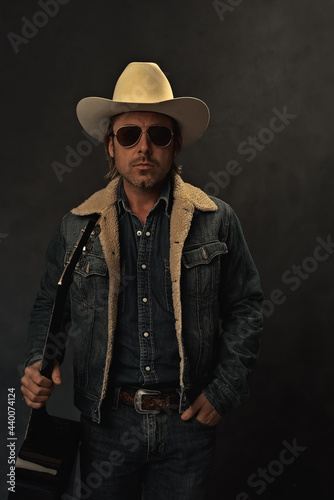 Man in denim jacket, white cowboy hat and aviator sunglasses with acoustic western guitar in front of a grey wall. © ysbrandcosijn