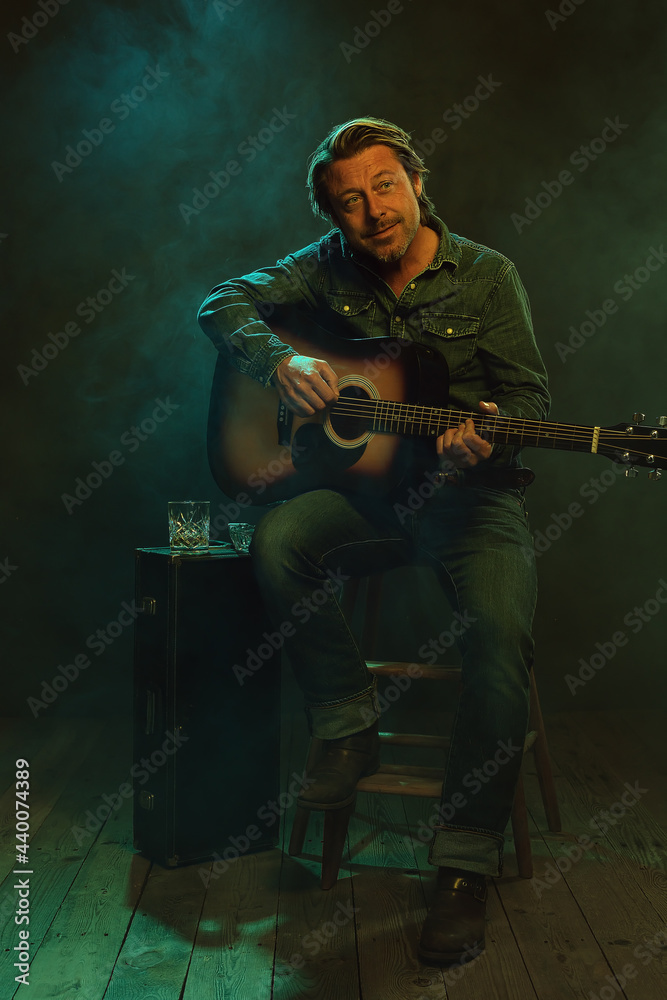Obraz premium Musician sitting on a wooden stool plays an acoustic guitar with next to him a guitar case with a glass of whiskey and an ashtray on it.