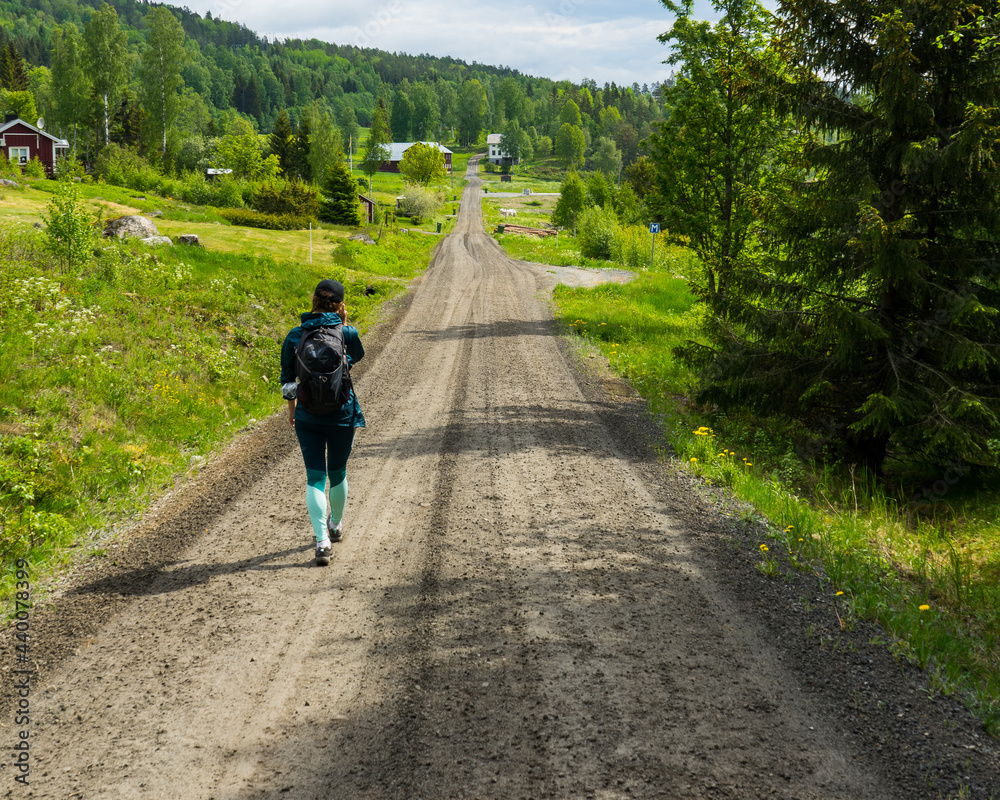 Girl walking down gravel road with green fields and forest scenery on the sides. High Coast area, northern Sweden