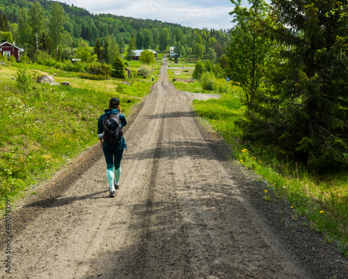 Girl walking down gravel road with green fields and forest scenery on the sides. High Coast area, northern Sweden