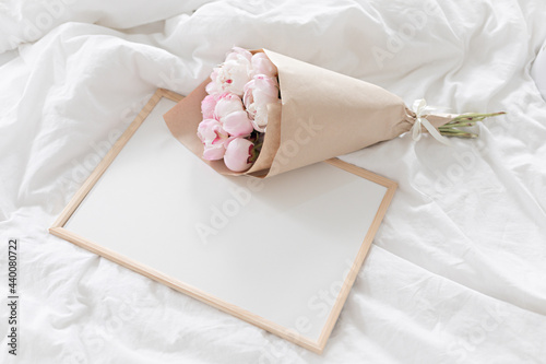White frame mockup on the bed. Bouquet of pink peonies in craft packaging. Scandivanavian white interior.