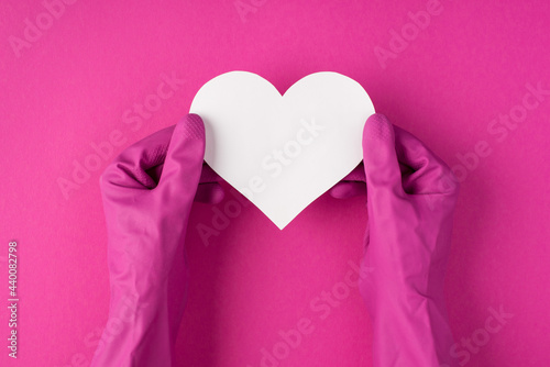 Top view photo of hands in pink gloves holding white paper heart on isolated pink background with blank space