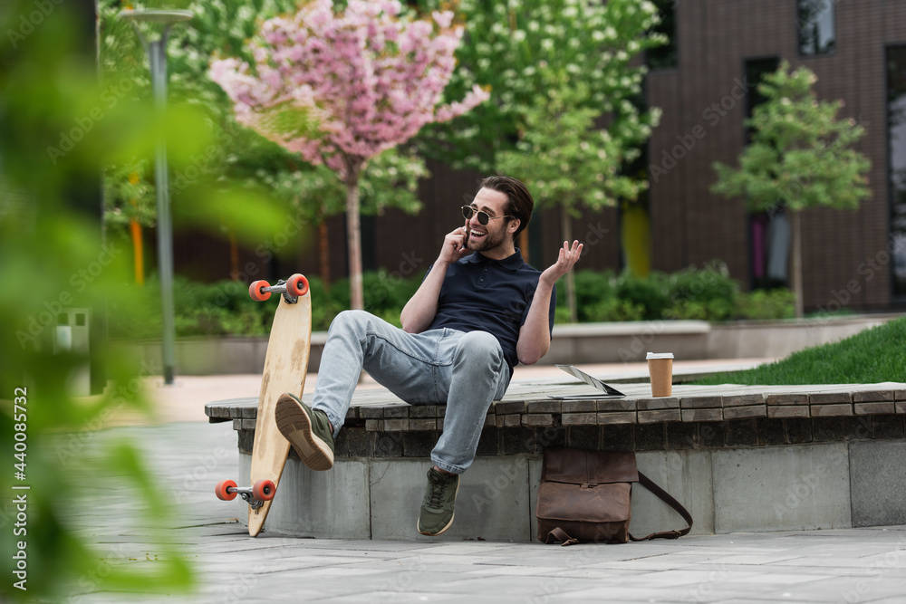 happy man in sunglasses talking on smartphone near longboard and gadgets. happy man in sunglasses talking on smartphone near longboard and gadgets.