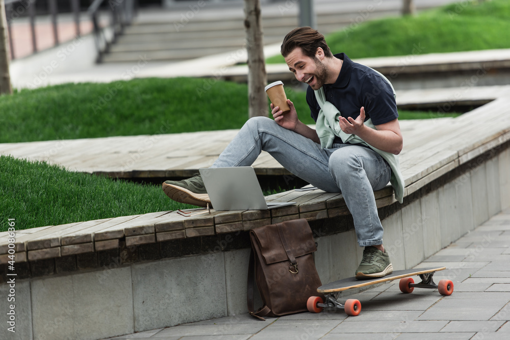 cheerful man in polo shirt and sweatshirt holding coffee to go and laughing while sitting near longboard and gadgets.