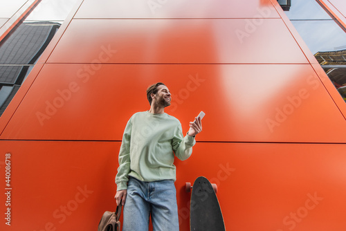 Quadro su tela low angle view of cheerful man in sweatshirt holding backpack and smartphone near longboard and orange wall