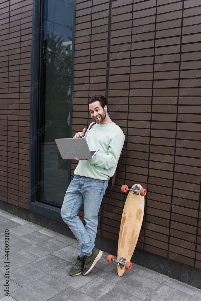 full length of happy freelancer in sweatshirt and wireless earphones using laptop outside.