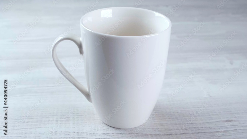 White mug on a light wooden table and the background of a modern room. Empty beverage cup for your design. You can place text, image and logo. White mug on a light wooden table and the background of a modern room. Empty beverage cup for your design. You can place text, image and logo.