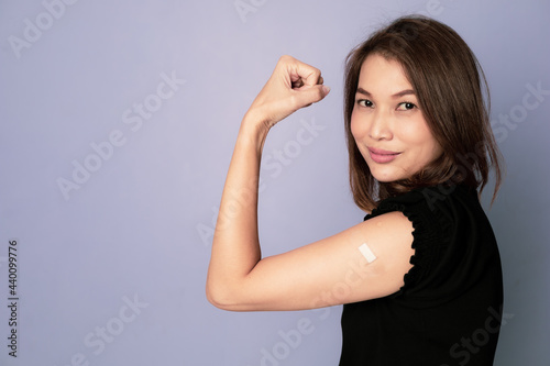 Portrait of vaccinated Asian woman showing plaster band aid on arm and show fist punch in winning and trust sign after coronavirus vaccine injection look at camera. Getting Vaccination Concept.