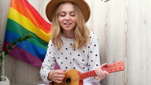 4k Young millennial hippie woman sitting on balcony play guitar. Music lesson and singer sitting. LGBTQ rainbow flag on background. Online music tuition. Playing instrument and enjoying songs 
