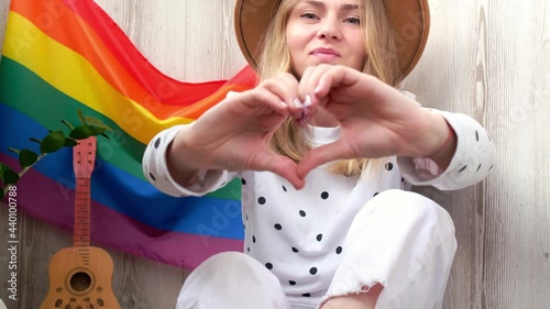 4k Young blonde millennial hippie woman showing heart sign with hands with rainbow LGBTQ flag at home balcony. Peace and freedom the symbol of LGBT, spending and sharing loving time. Support LGBTQ 