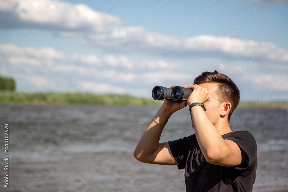 Young guy with a fitness bracelet looks through binoculars against the background of the river
