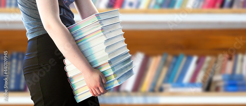 Canvas Print Girl holding a huge pile of books in the library, panoramic view, copy space for text