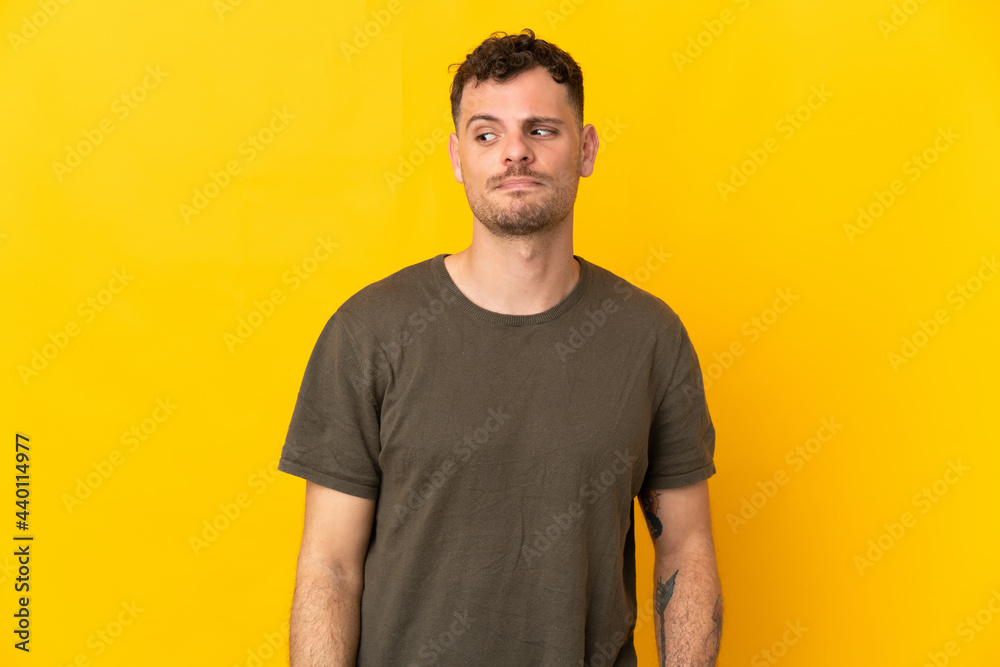 Young caucasian handsome man isolated on yellow background having doubts while looking side Young caucasian handsome man isolated on yellow background having doubts while looking side