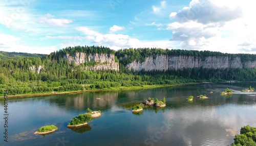 A huge rock mass stands like a wall along the river . Beautiful blue sky and shaggy clouds . Green forest along the river, small islands . Catamarans are rafted on the river .