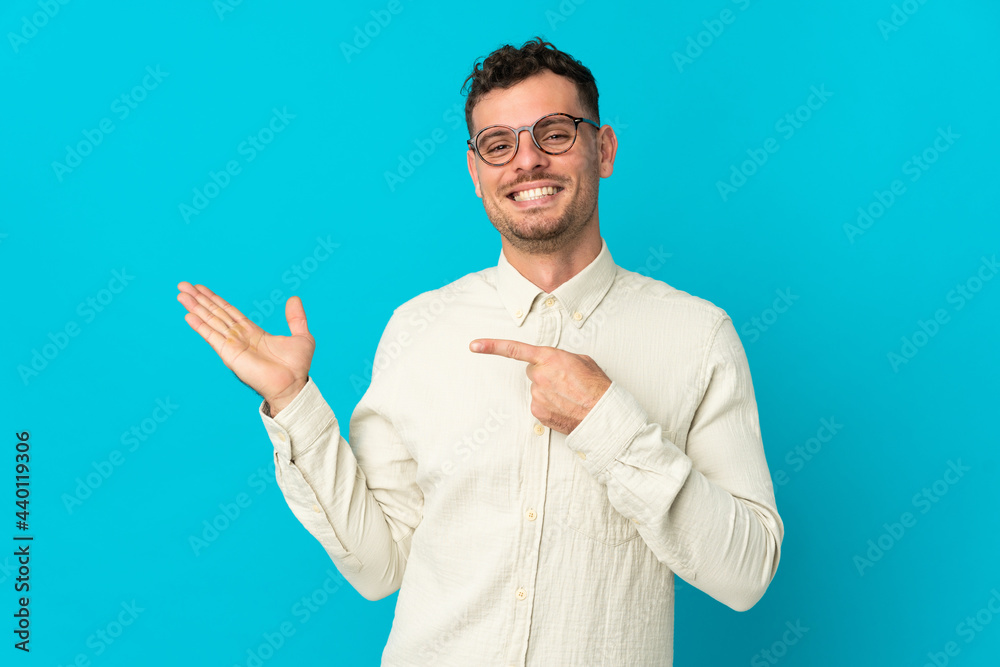 Young caucasian handsome man isolated on blue background holding copyspace imaginary on the palm to insert an ad Young caucasian handsome man isolated on blue background holding copyspace imaginary on the palm to insert an ad