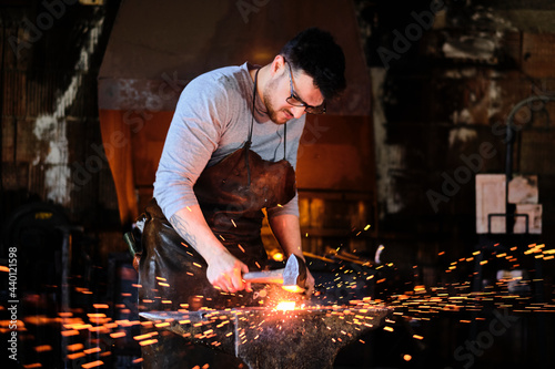 Young male blacksmith wearing eyeglasses forging with hammer at workshop
