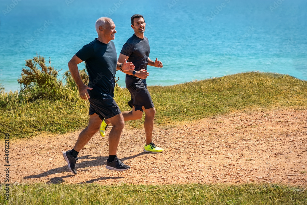 Obraz premium Fitness instructor gives instructions to an elderly man, a retired client, jogging on the seashore, endurance training.