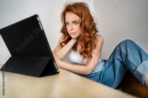 Beautiful redhead woman using digital tablet at table against white wall in room