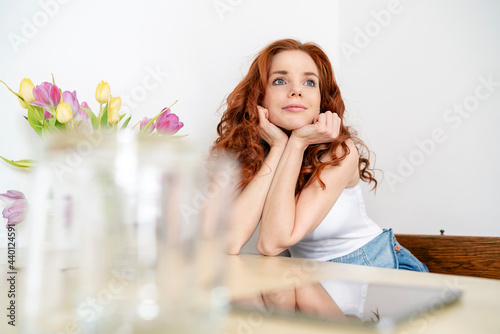 Thoughtful redhead mid adult woman sitting with hands on chin while leaning on table at home