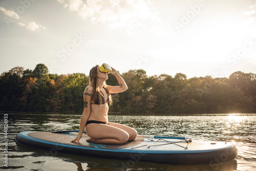 Young woman with tatto kneeling on paddleboard, using VR goggles
