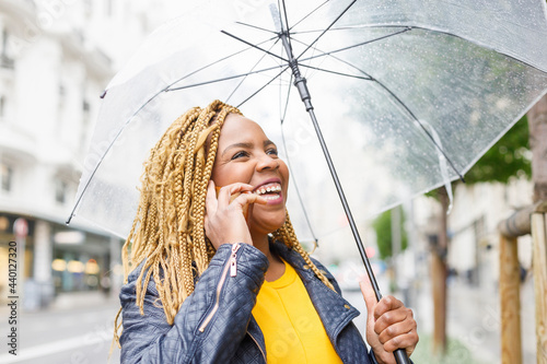 Cheerful woman with braided hair talking on phone while holding umbrella