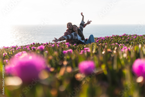 Young couple with arms outstretched sitting at flower filed by sea