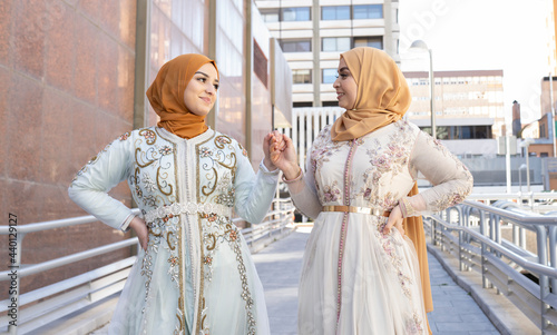 Friends wearing hijab and dress smiling while holding hands on bridge photo