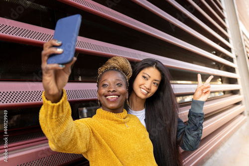 Young woman taking selfie with female friend showing peace sign by wall photo